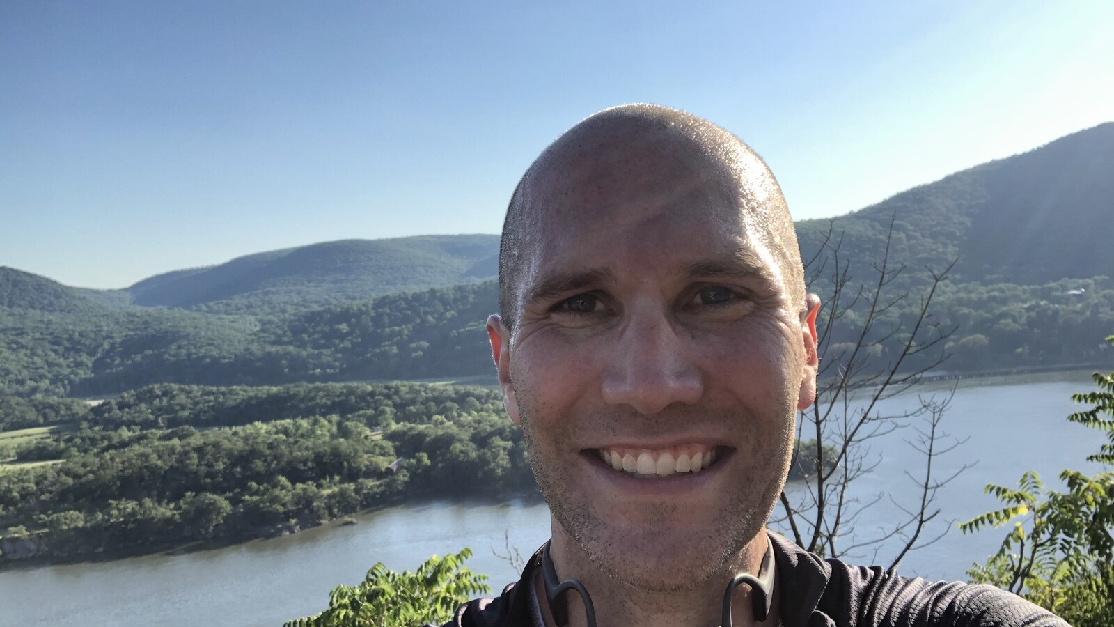 Chris smiling after a summer bike ride with the Hudson River and mountains behind him