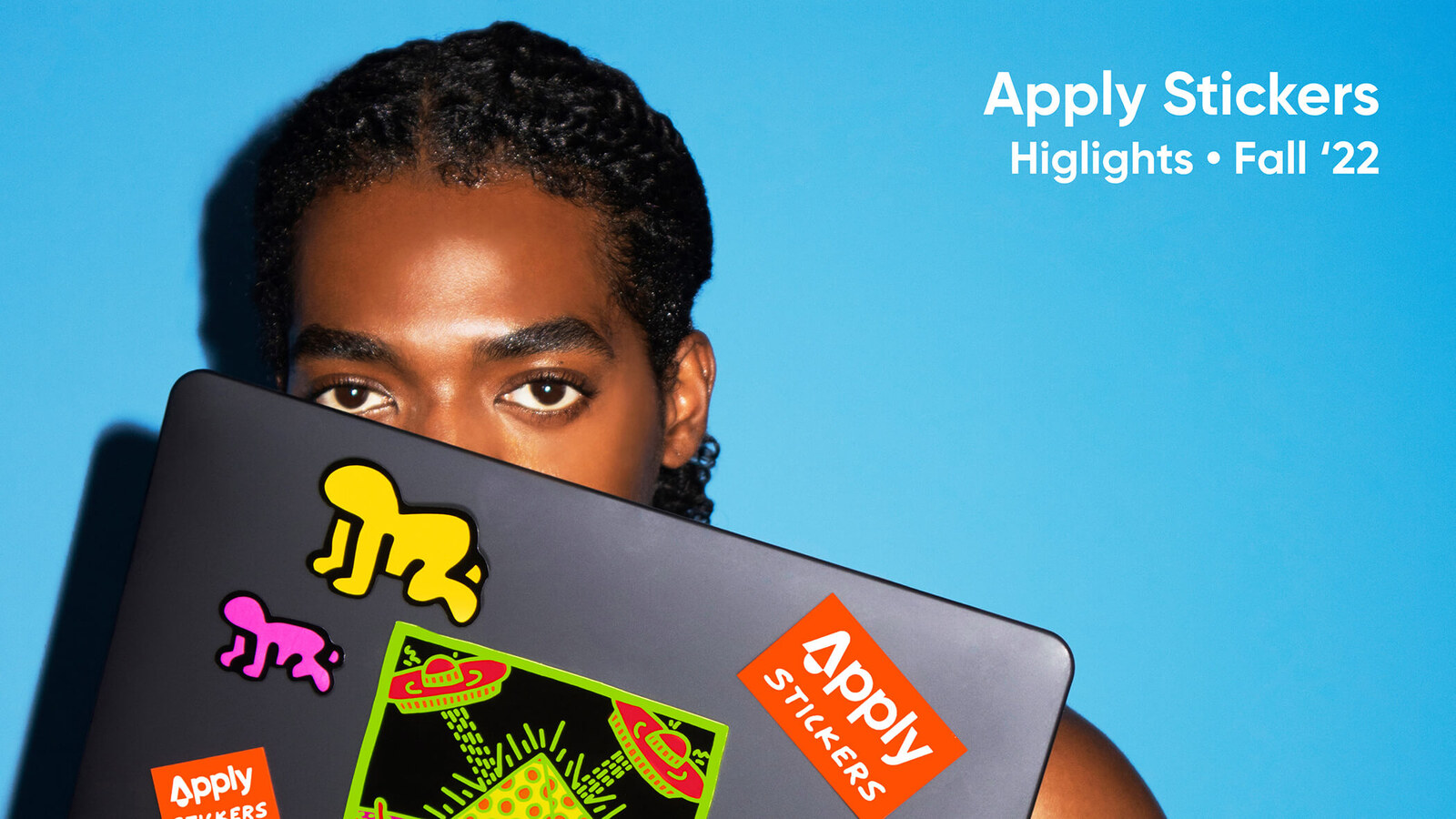 A model holding a laptop covered in colorful Apply Stickers against a blue backdrop