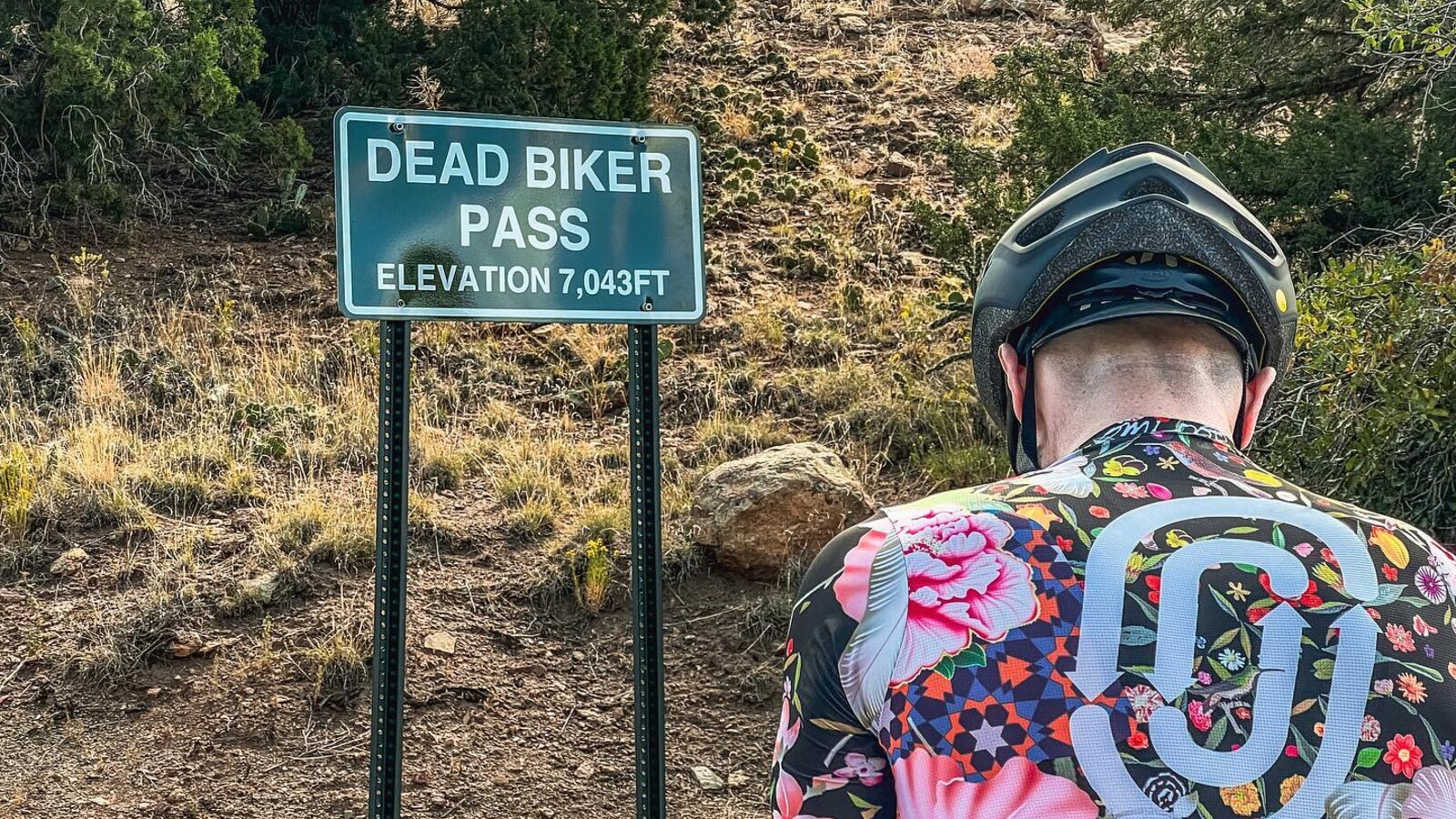 A cyclist in a flowered jersey stopped beside the Dead Biker Pass sign at 7,043 feet
