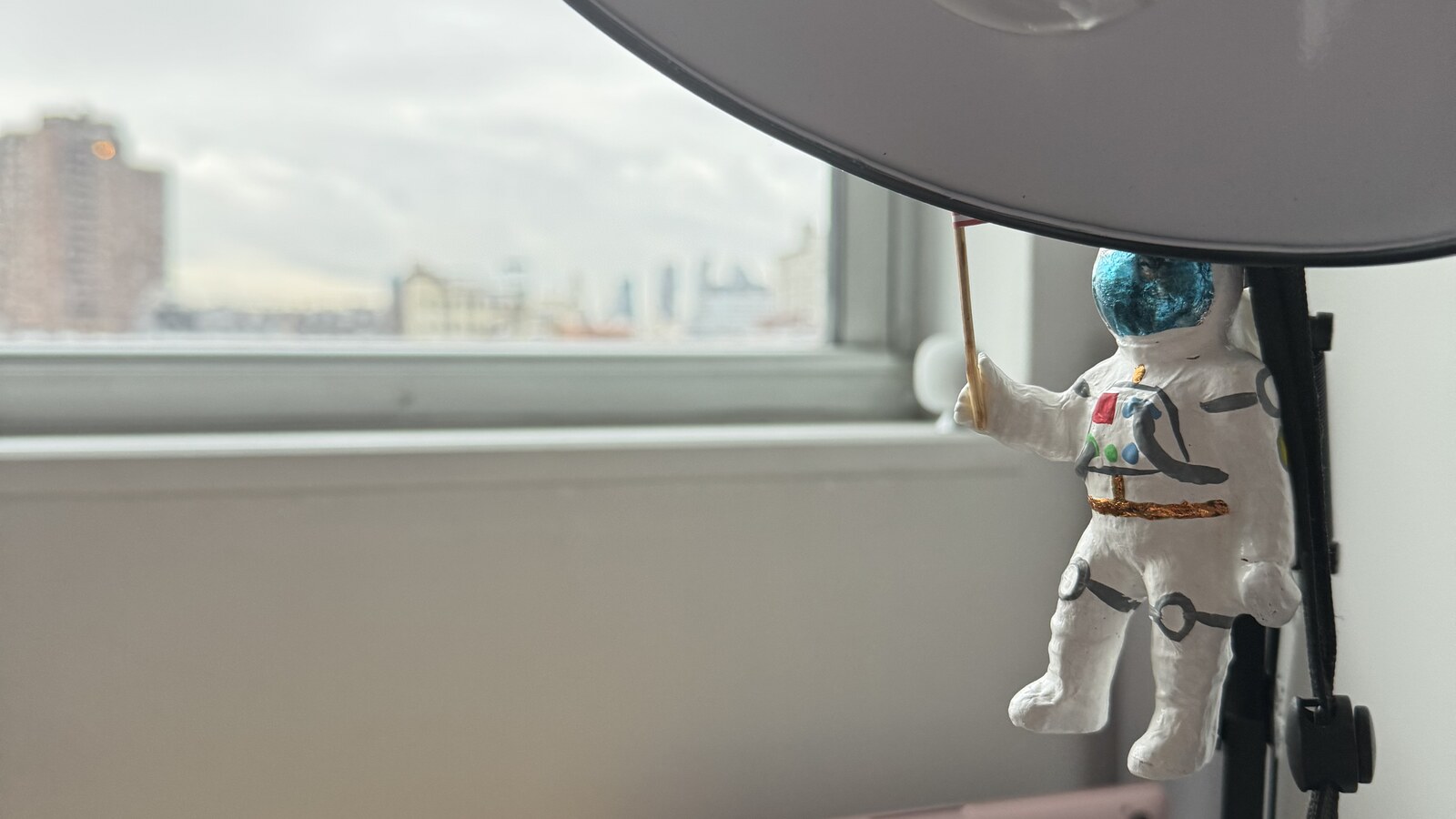 A small astronaut figure hanging beneath a large desk lamp beside a Williamsburg apartment window